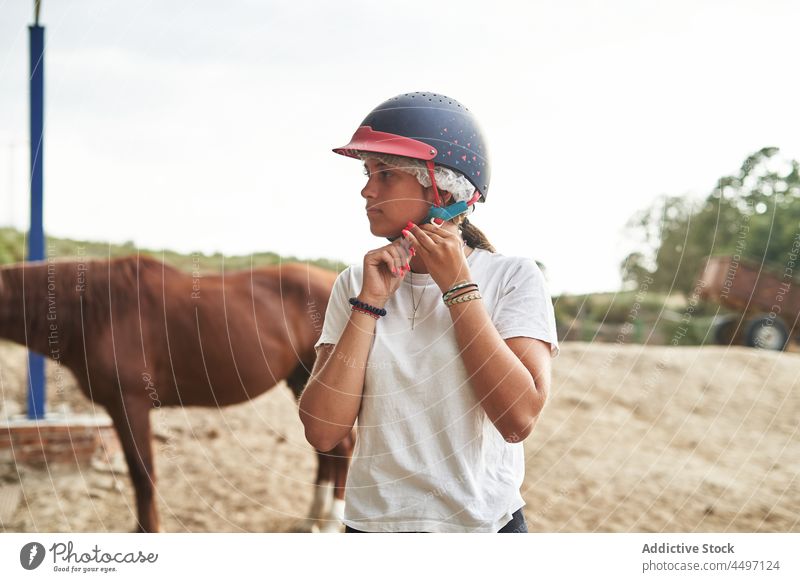 Mädchen setzt Jockeymütze in der Nähe von Pferden auf einer Ranch auf Teenager Tier angezogen Pferdestall Bauernhof Schutzhelm Landschaft stehen tagsüber