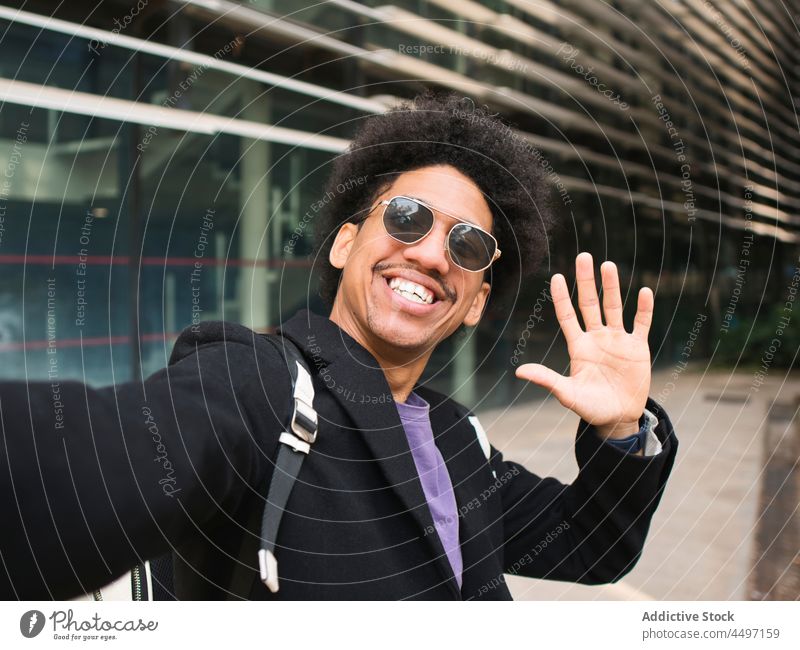 Fröhlicher schwarzer Mann lächelt fröhlich beim Selfie machen Lächeln heiter Wellenhand trendy selbstsicher Stil Optimist Großstadt selbstbewusst modern