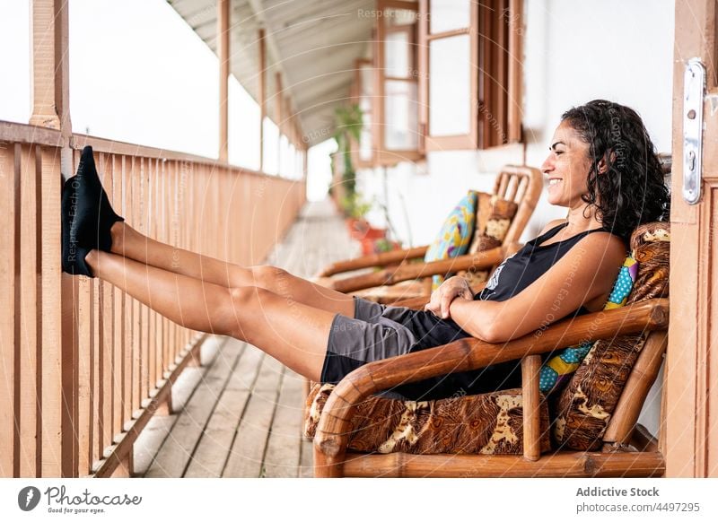 Glückliche ethnische Frau, die sich auf der Terrasse ausruht und lächelt ruhen Lächeln heiter Urlaub Tourist Haus Feiertag Armsessel genießen Ausflug jung
