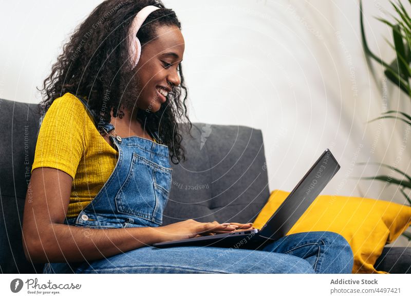 Glückliche afroamerikanische Frau mit Laptop im Wohnzimmer Lächeln benutzend Kopfhörer Musik Stock Sofa Freude Jeansstoff heimwärts zuhören lässig sitzen