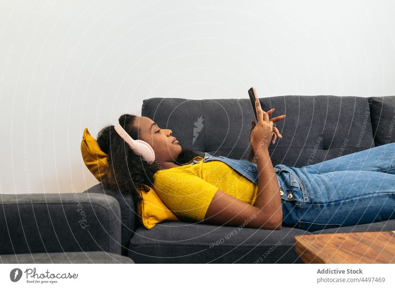 Schwarze Frau mit Kopfhörern am Smartphone im Zimmer krause Haare Browsen Lügen positiv Blogger zuhören Musik Textnachricht Afroamerikaner zuschauen schwarz