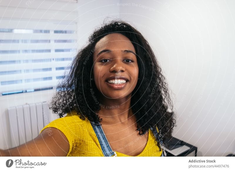Lächelnde schwarze Frau, die in einem hellen Raum ein Selfie macht Smartphone heiter positiv Apparatur Gerät fotografieren Blogger Afroamerikaner krause Haare