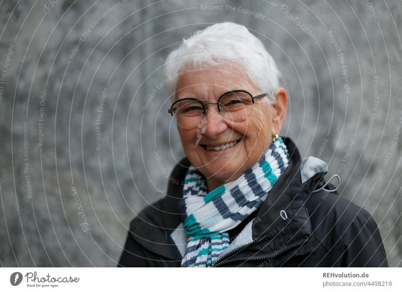 Portrait einer Seniorin vor Beton Oberkörper grauhaarig weißhaarig authentisch Erwachsene glücklich Glück Zufriedenheit Lächeln natürlich 60 und älter