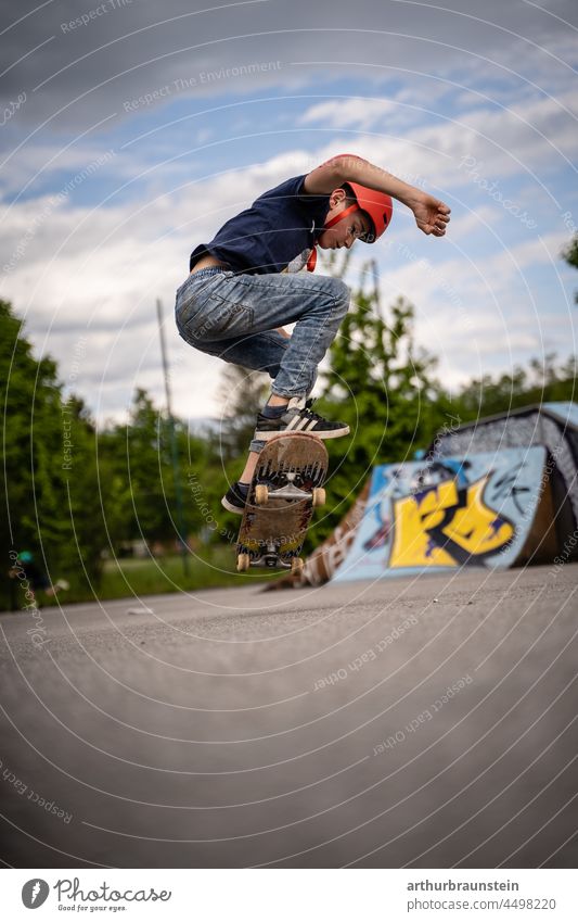 Junge in Jeans und mit Helm macht einen Ollie im Skatepark im Freien Tageslicht asphalt bewegung bewölkt board boardsport cool deck draussen freizeit grafitti