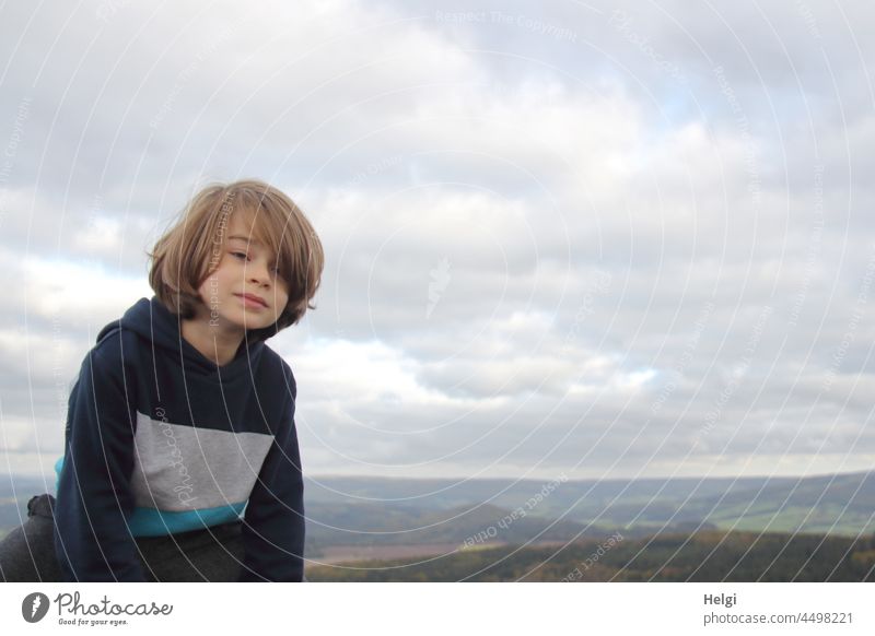 Junge sitzt in der Natur vor bewölktem Himmel, im Hintergrund Berge der Rhön Kind Mensch draußen sitzen schauen Porträt Oberkörper Wolken Ausflug Außenaufnahme
