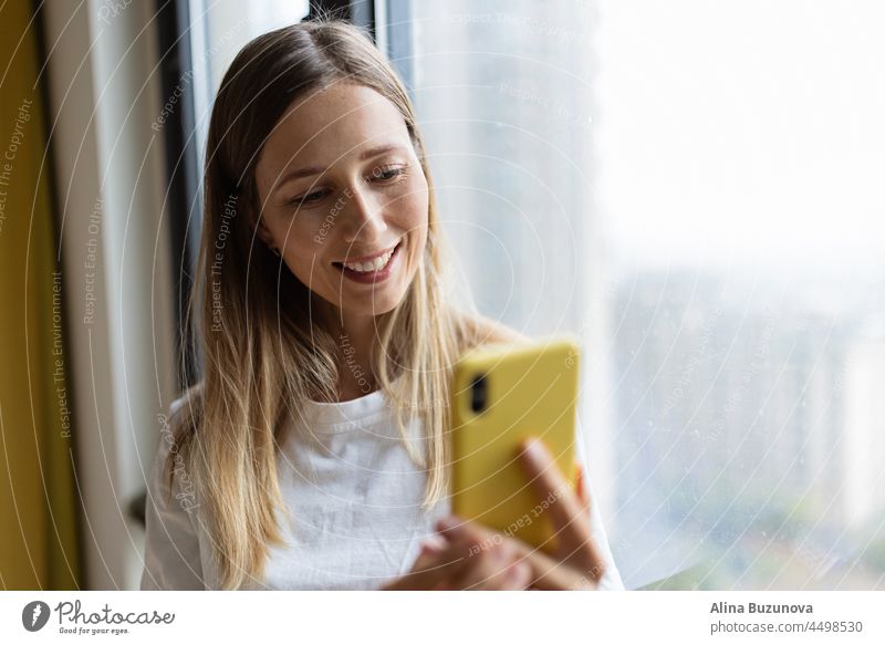 Junge hübsche Millennial-Frau hält Handy zu Hause oder in einem Café in der Nähe des Fensters. Video-Chat Lebensmittel-Lieferung Online-Shopping