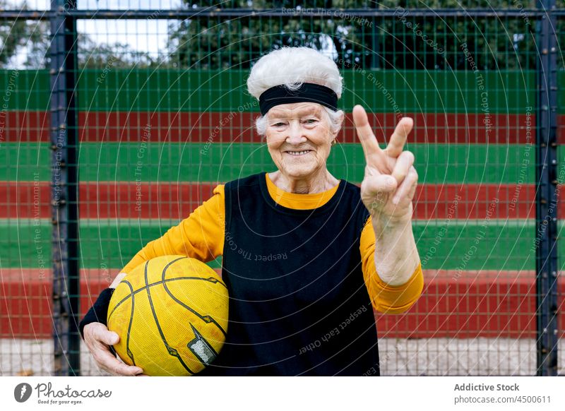 Positive Seniorin mit Ball auf Spielplatz Frau Basketball spielen Stirnband Sportpark zwei Finger Zeichen Frieden Sieg Gesunder Lebensstil Hobby Training reif