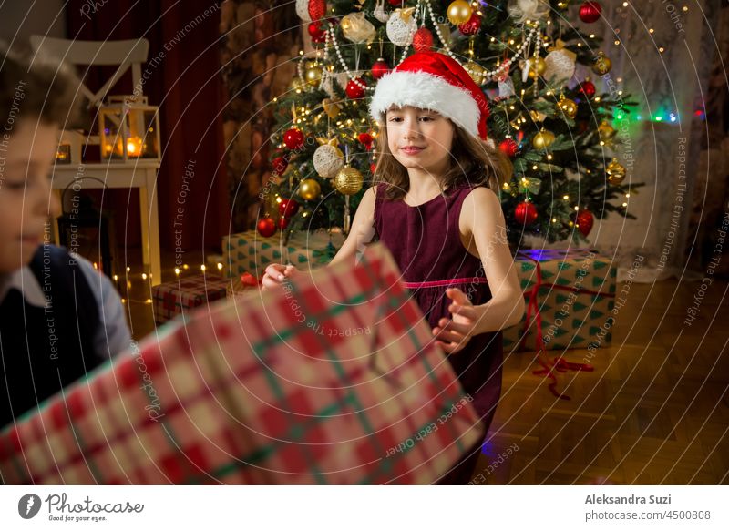 Aufgeregte niedliche Kinder, die fröhlich lächelnd Weihnachtsgeschenke öffnen. Schön dekoriert Wohnzimmer mit Lichtern und Weihnachtsbaum. Kinder haben Spaß, feiern Familie Urlaub