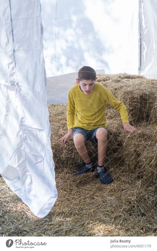 Kleiner Junge mit Autismus sitzt auf Heuballen bei einem Herbstfest; sensorisches Spiel und Erkundung fallen Ballen Zelt Kürbispflaster Bauernhof Stadtfest