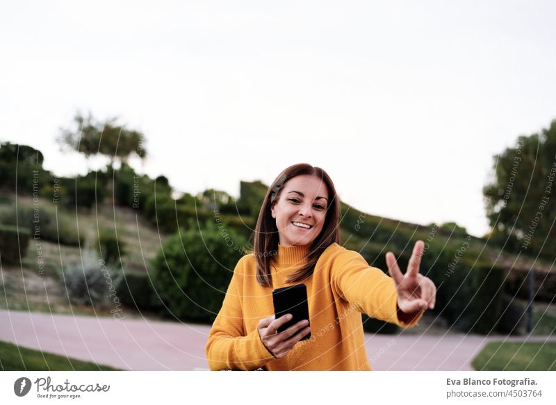 Unbekümmerte kaukasische Frau, die ein V-Zeichen macht, einen gelben Pullover trägt und ein Mobiltelefon im Freien im Park bei Sonnenuntergang benutzt. Technologie und Lebensstil