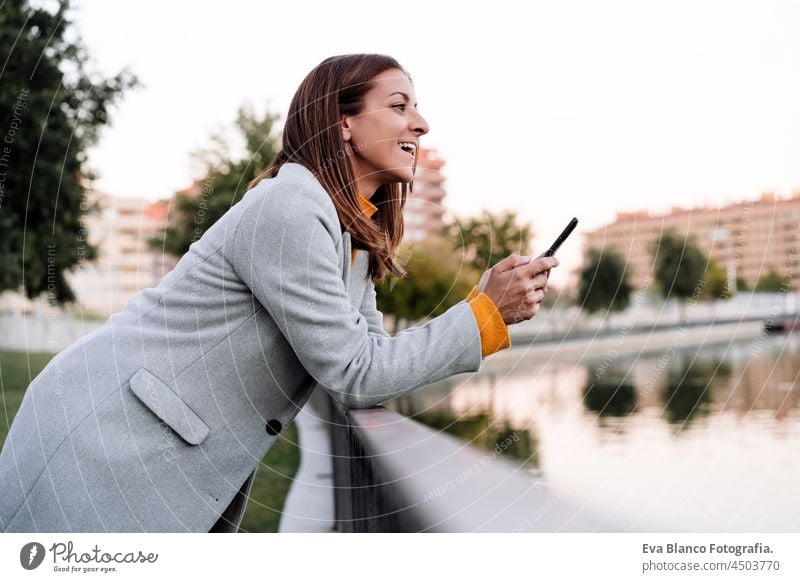 Lächelnde kaukasische Frau mit gelbem Pullover und grauem Mantel, die ein Mobiltelefon im Freien im Park bei Sonnenuntergang benutzt. Technologie und Lebensstil