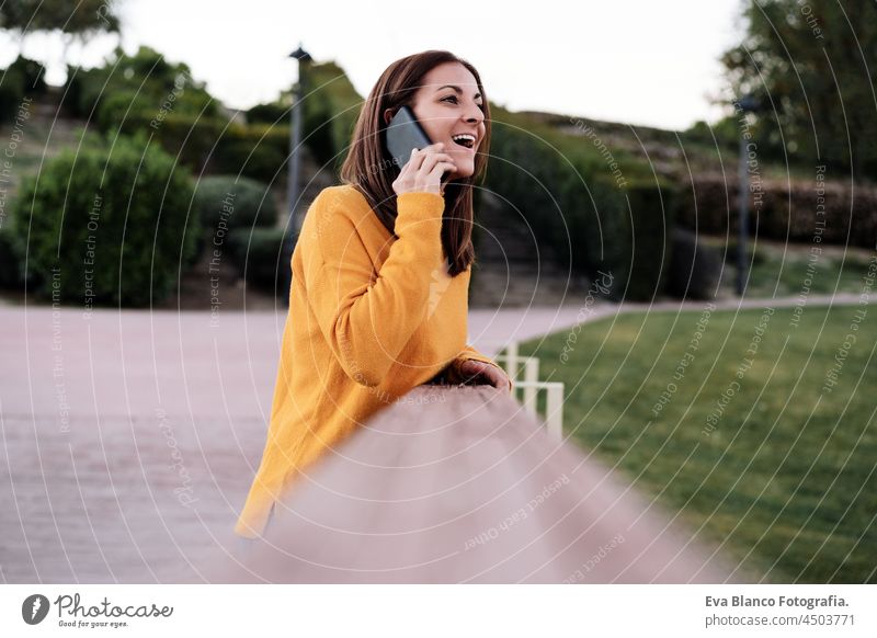 glückliche kaukasische Frau im gelben Pullover, die im Freien im Park bei Sonnenuntergang mit ihrem Handy telefoniert. Technologie und Lebensstil Sprechen