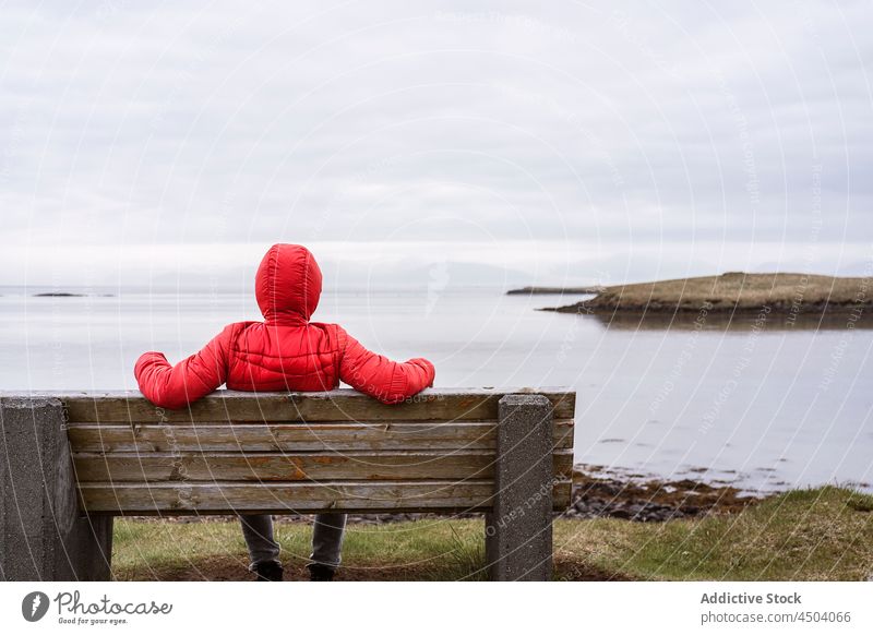 Anonyme Frau sitzt auf einer Bank und bewundert das Meer an einem bewölkten Tag Landschaft Ufer Oberbekleidung Natur bewundern Wasser Küste Island