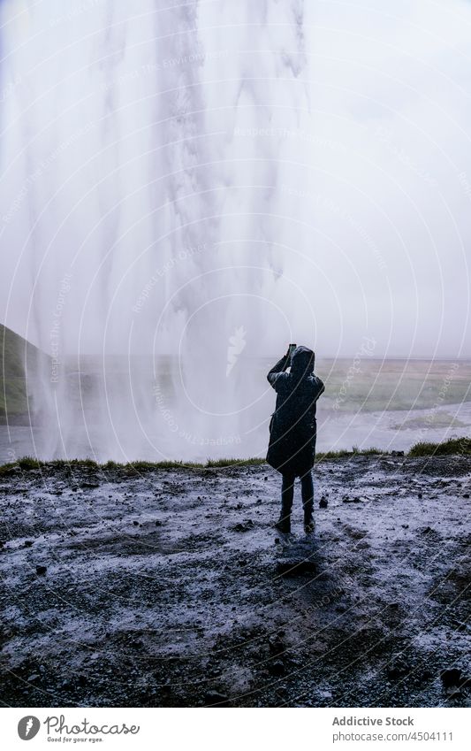 Anonymer Tourist, der an einem bewölkten Tag mit seinem Smartphone einen beeindruckenden Wasserfall fotografiert Person fotografieren Natur Ausflug erkunden