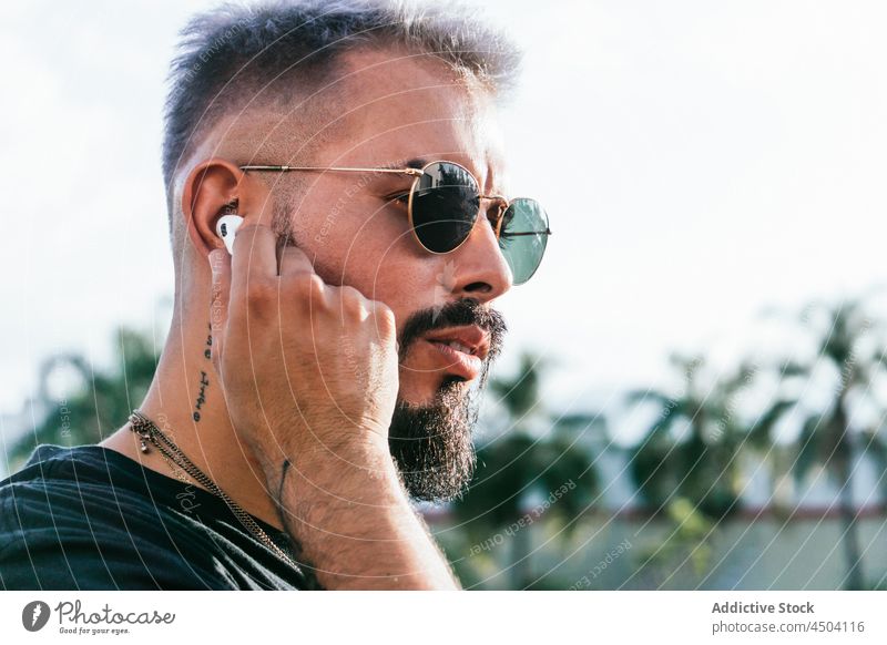 Seriöser Mann mit Kopfhörern auf der Straße bei Tag Tattoo Sonnenbrille Vollbart besinnlich trendy zuhören anhaben Musik männlich nachdenklich Baum urban Stil