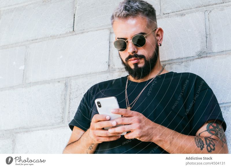 Fröhlicher Mann mit Tattoos, der ein Smartphone benutzt Nachricht Mobile benutzend männlich Sonnenbrille Apparatur Gerät Straße urban Typ Tageslicht
