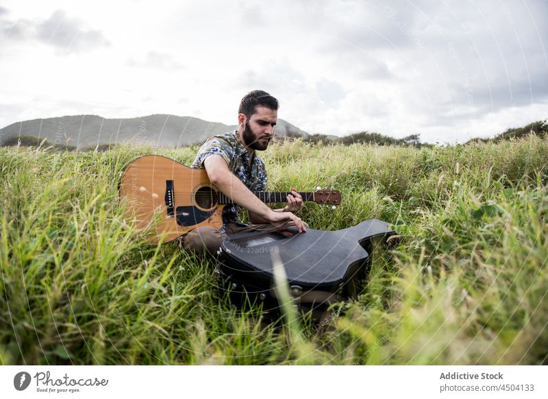 Gitarrist sitzt auf einem Feld gegen einen Hügel in der Landschaft Mann Musiker akustisch Gitarre Gras Instrument Natur männlich wolkig Windstille sitzen