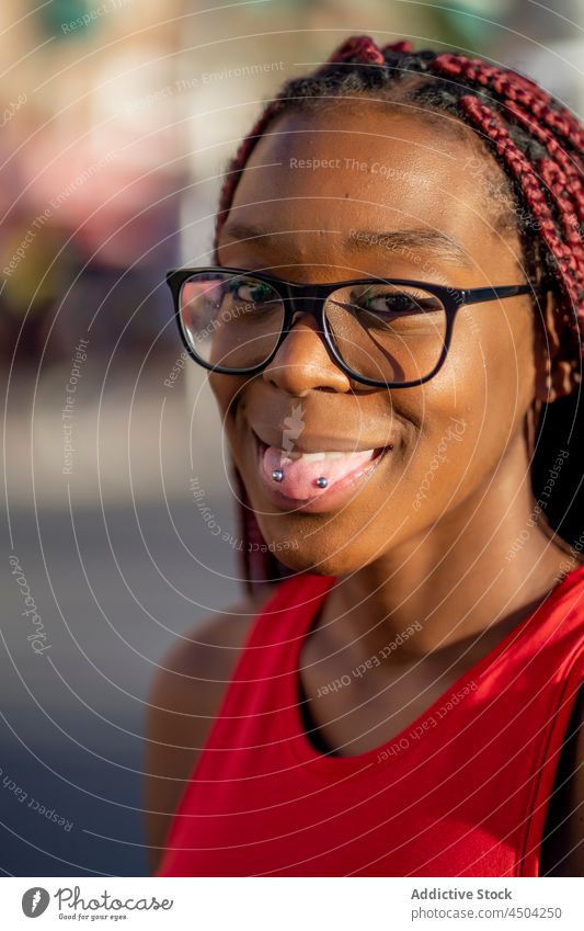 Fröhliche schwarze Frau zeigt Zunge mit Piercing im Sonnenlicht Geflecht positiv Straße froh ausspannen Stil trendy Optimist Afroamerikaner Glück Vorschein
