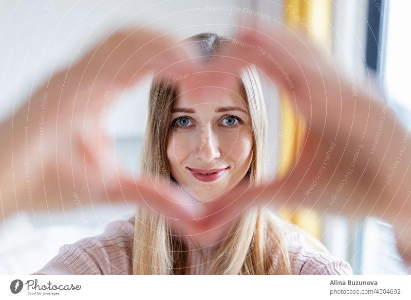 Nahaufnahme lächelnde attraktive junge kaukasische Frau mit blondem Haar, 30-35 Jahre alt, zeigt herzförmige Geste zu Hause während der Coronavirus-Covid-19-Pandemie-Quarantäne und Selbstisolierung