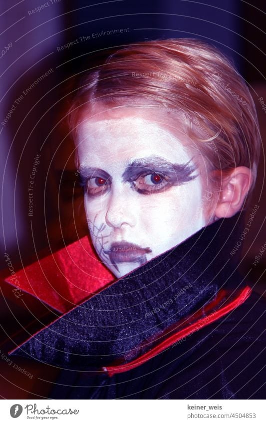 Ein als Vampir geschminktes Kind Kostüm Fasching Karneval Kindheit Dracula Halloween rote Augen Mantel Kragen Mantelkragen Gesicht weiß blond Porträt Farbfoto