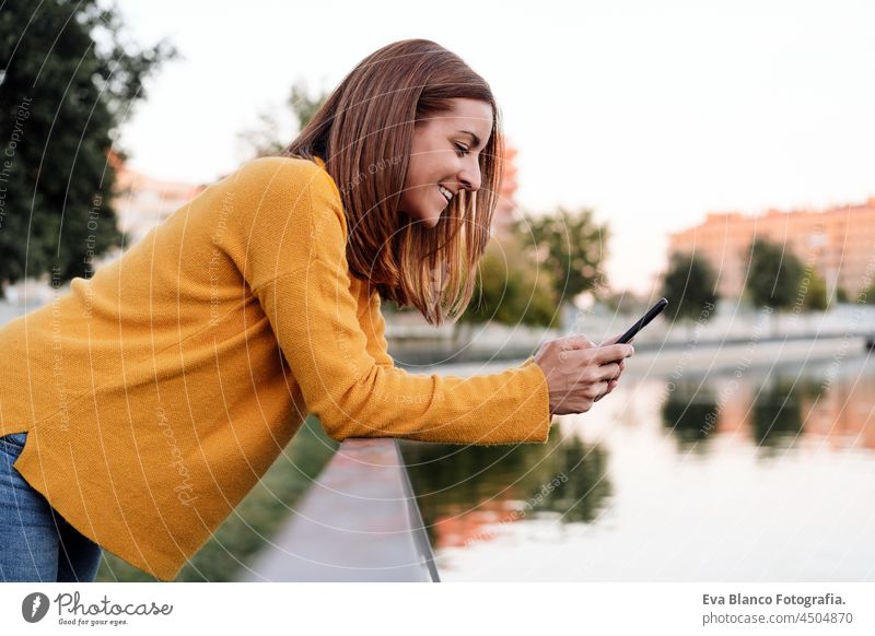 Lächelnde kaukasische Frau mit gelbem Pullover, die ein Mobiltelefon im Freien im Park bei Sonnenuntergang benutzt. Technologie und Lebensstil Handy Sprechen
