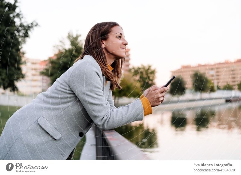 Lächelnde kaukasische Frau mit gelbem Pullover und grauem Mantel, die ein Mobiltelefon im Freien im Park bei Sonnenuntergang benutzt. Technologie und Lebensstil