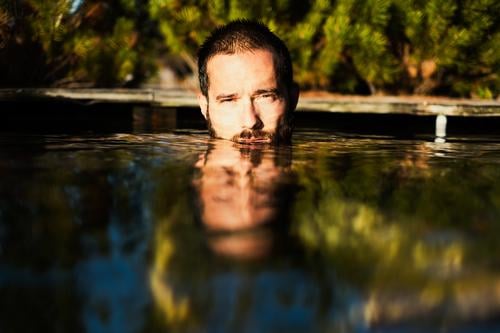 Ein Mann, das Gesicht halb unter Wasser, badet draußen, umgeben vom Natur, bei Sonnenschein baden Sommer See Spiegelumg Portrait Mensch Wärme Sonnenlicht