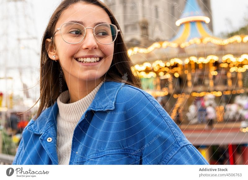 Lächelnde Frau in der Nähe eines Karussells im Stadtpark Reling Großstadt Straße positiv Gebäude Brille sich auf die Hand lehnen Lifestyle jung Metall stehen