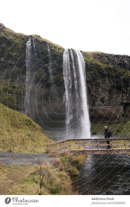 Anonymer Mann, der einen Wasserfall beobachtet, der von einer Felsklippe in Island herabstürzt Reisender Klippe Steg bewundern Schlucht Natur erkunden Teich