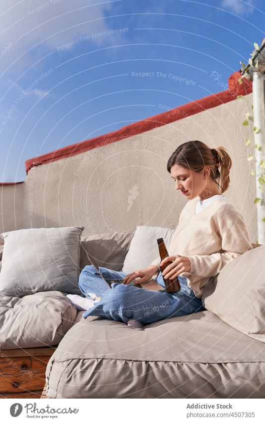 Frau mit Bierflasche beim Stöbern im Laptop benutzend Kälte Internet Dachterrasse online Tippen Terrasse zuschauen Gerät Netbook Browsen Sofa modern Alkohol