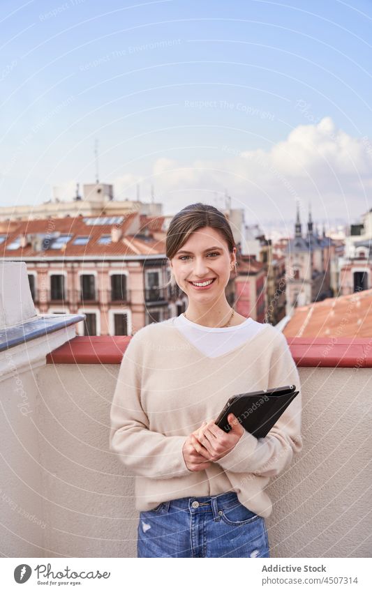 Lächelnde Frau mit Tablet auf einem Balkon stehend benutzend Internet Vorschein Dachterrasse positiv online charmant Telefon Terrasse lässig Jeanshose Optimist
