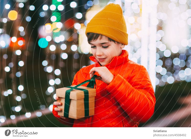 Porträt eines glücklichen Jungen, der auf der Abendstraße steht und ein Weihnachtsgeschenk öffnet. Weihnachtsgeschenke, Urlaubsatmosphäre. Das Konzept von Weihnachten und Neujahr