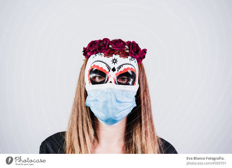 Frau trägt mexikanische Gesichtsmaske während Halloween-Feier. Skelett-Kostüm und rote Rosen Diadem auf dem Kopf. Frau trägt blaue Gesichtsmaske während Pandemie Corona-Virus. Halloween-Party-Konzept