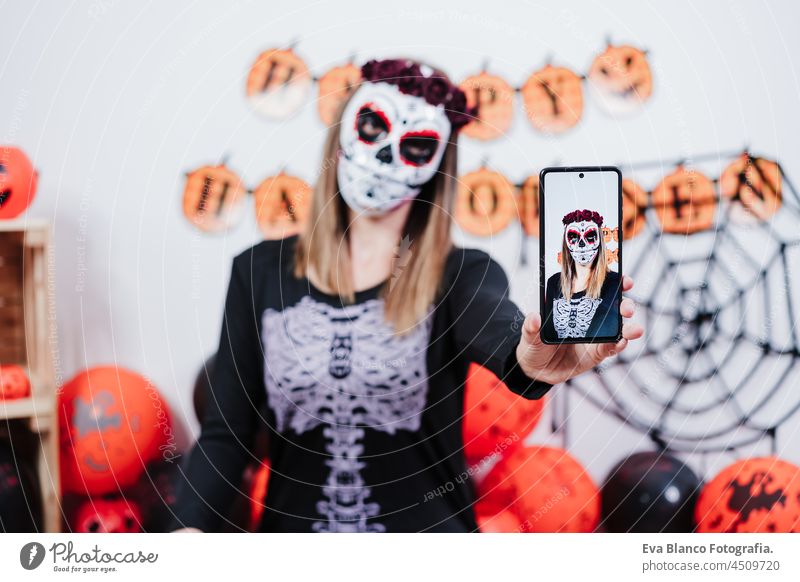 Frau trägt mexikanische Gesichtsmaske während Halloween-Feier. Aufnahme selfie mit Handy. Frau trägt Skelett-Kostüm und rote Rosen Diadem auf dem Kopf. Halloween-Party-Konzept
