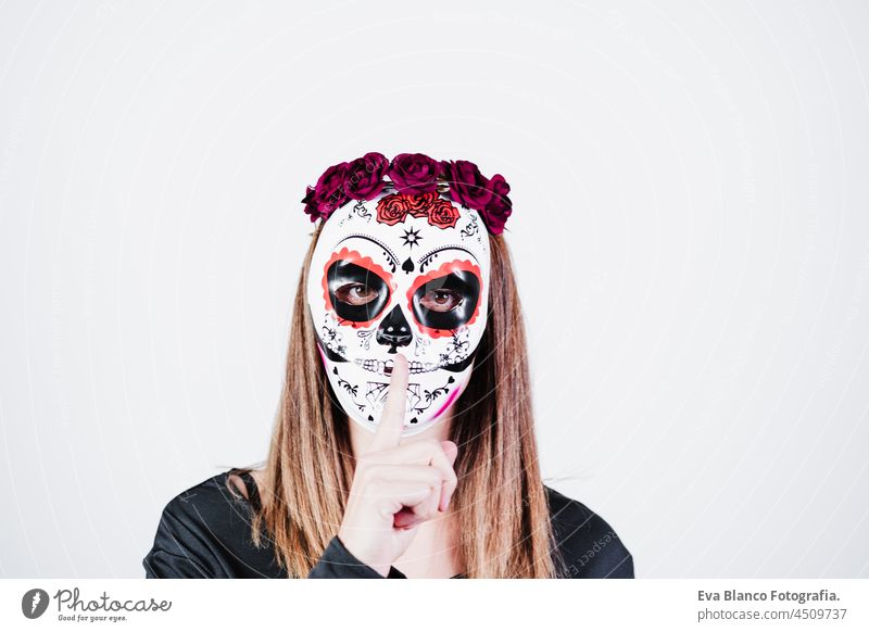 Frau trägt mexikanische Gesichtsmaske während Halloween-Feier. Skelett-Kostüm und rote Rosen Diadem auf dem Kopf. Frau macht Stille Geste mit dem Finger. Halloween-Party-Konzept