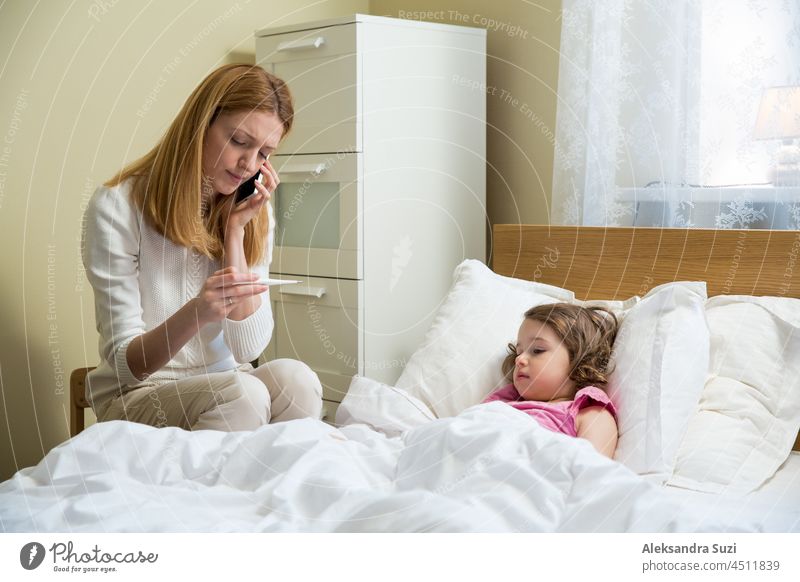 Mutter misst die Temperatur ihres kranken Kindes. Krankes Kind mit hohem Fieber im Bett liegend und Mutter hält Thermometer. Hand auf der Stirn. Coronavirus