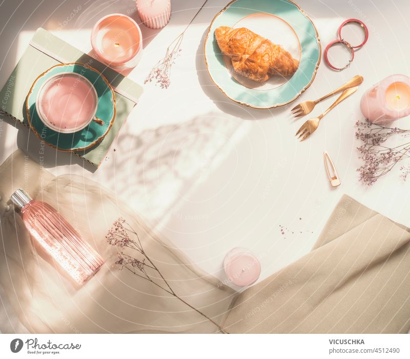 Moderne Morgenroutine mit Croissant, Blumen Tee, Duftkerzen, Gabeln, rosa Parfüm und Notizbuch auf weißem Tisch. Romantischer femininer Hintergrund. Draufsicht mit Kopierbereich.