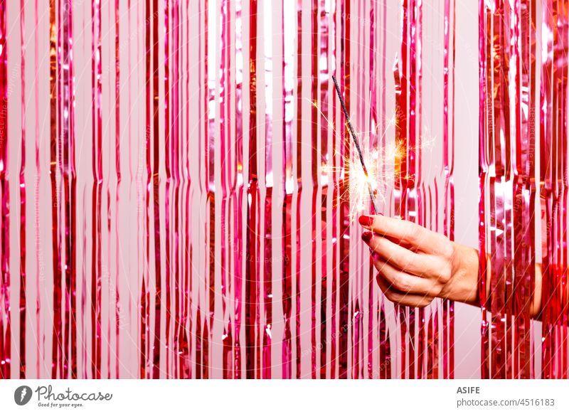 Frauenhand, die mit einem brennenden Sarkler ein rosa Lametta durchbricht. Wunderkerze Hand Party Gardine Neujahr Folie Glitter schimmern retro rot glühen Glanz