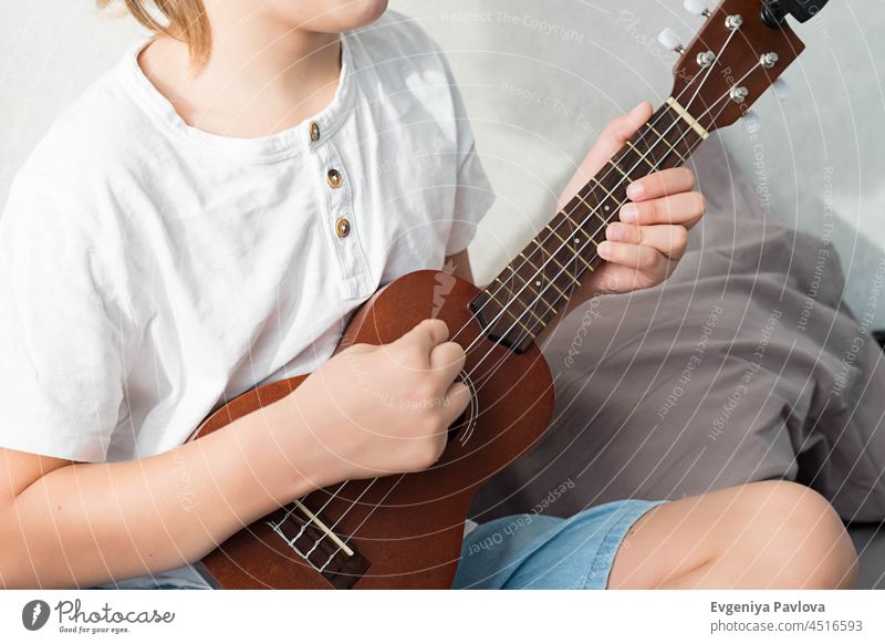 Junger Junge stimmt Ukulele zu Hause. Blondhaariger Junge sitzt auf der Couch und spielt akustische Gitarre. jung Spielen Tuning heiter Hobby Menschen Gesang