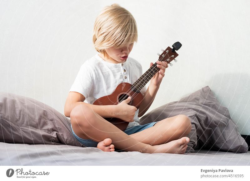 Junger Junge stimmt Ukulele zu Hause. Blondhaariger Junge sitzt auf der Couch und spielt akustische Gitarre. jung Spielen Tuning heiter Hobby Menschen Gesang