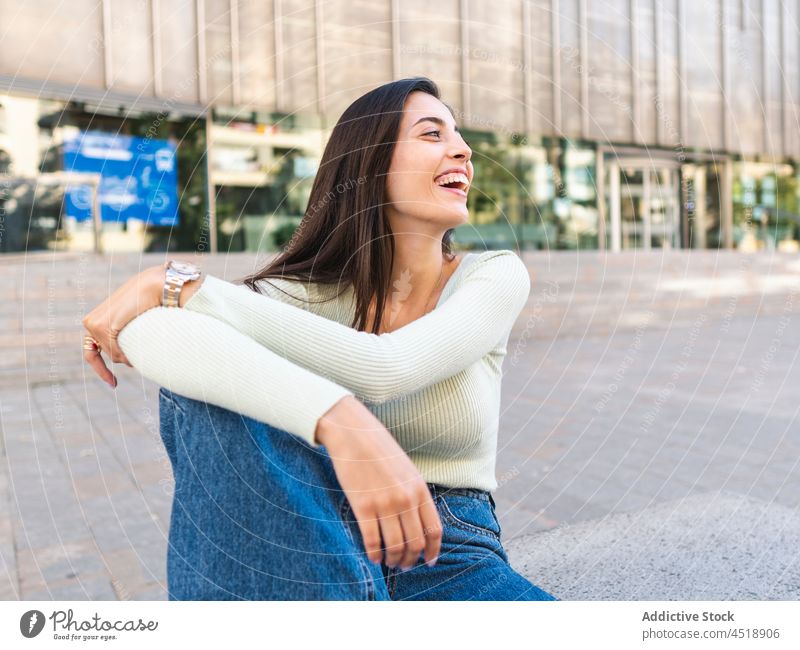 Positive Frau, die tagsüber auf der Straße auf einem Steinrand sitzt Großstadt urban Lächeln sitzen Glück Knie umarmend heiter Stil positiv charmant Stadt