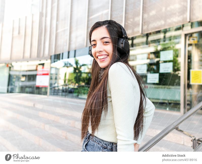 Glückliche Frau mit Kopfhörern auf der Straße stehend träumen Musik Windstille Großstadt Vorschein Stil urban Gebäude ausrichten braune Haare charmant Outfit