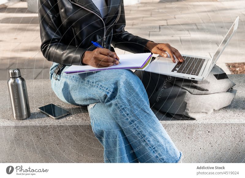 Anonyme schwarze Frau, die auf einem Laptop surft und in ein Notizbuch schreibt Browsen Straße online Internet schreiben Notebook Netbook Information Apparatur