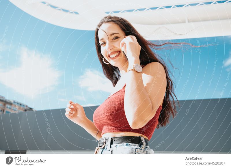 Junge arabische Frau, die einen Anruf tätigt, während sie Einkaufstaschen hält, Einkaufstag, modernes Outfit-Styling, Lächeln in die Kamera an einem sonnigen Tag