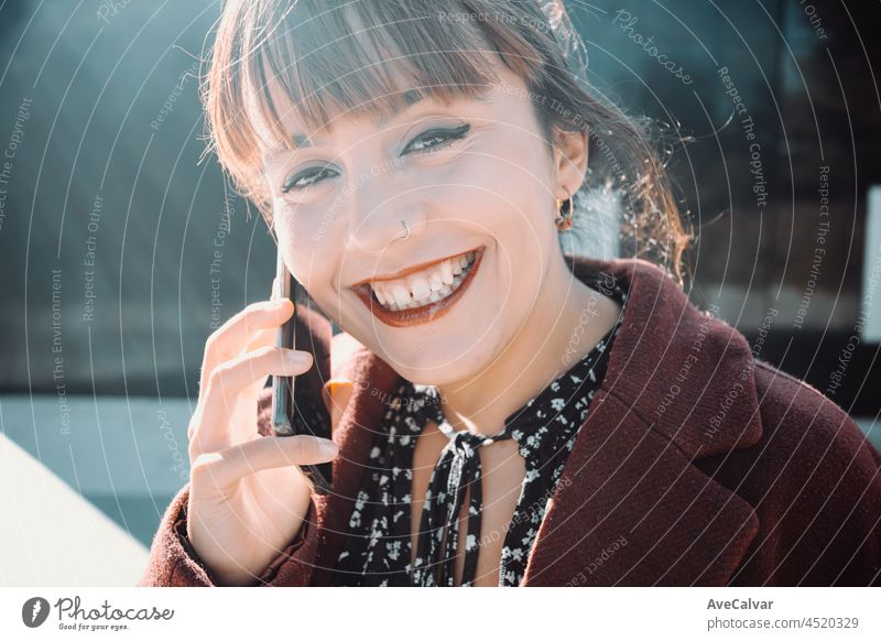 Junge Unternehmerin in einem gestylten Mantel, die in die Kamera lächelt, während sie bei Sonnenuntergang einen geschäftlichen Anruf tätigt, junges Unternehmen und zukünftiges Unternehmen
