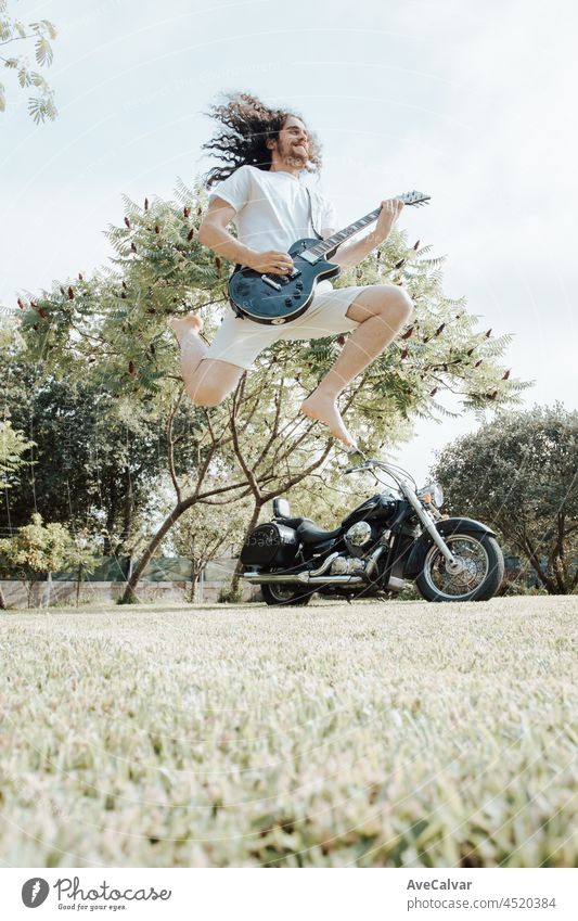 Ein Mann springt, während er eine E-Gitarre mit einem Motorrad als Hintergrund spielt. Heavy-Metal-Lifestyle-Konzept. Entspannter und sonniger Tag im Park.