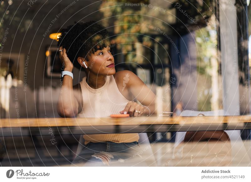 Selbstbewusste junge ethnische Frau schaut weg, während sie sich in einem Café ausruht Haare berühren ruhen trendy verträumt Smartphone Freizeit selbstbewusst