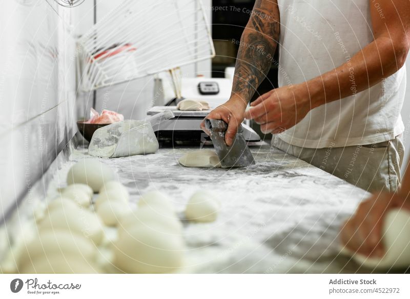 Crop-Bäcker schneiden Teig in der Küche Mann geschnitten Teigwaren Bäckerei Arbeit Zusammensein Prozess Mehl Brot roh Spielfigur kneten männlich Tattoo lässig