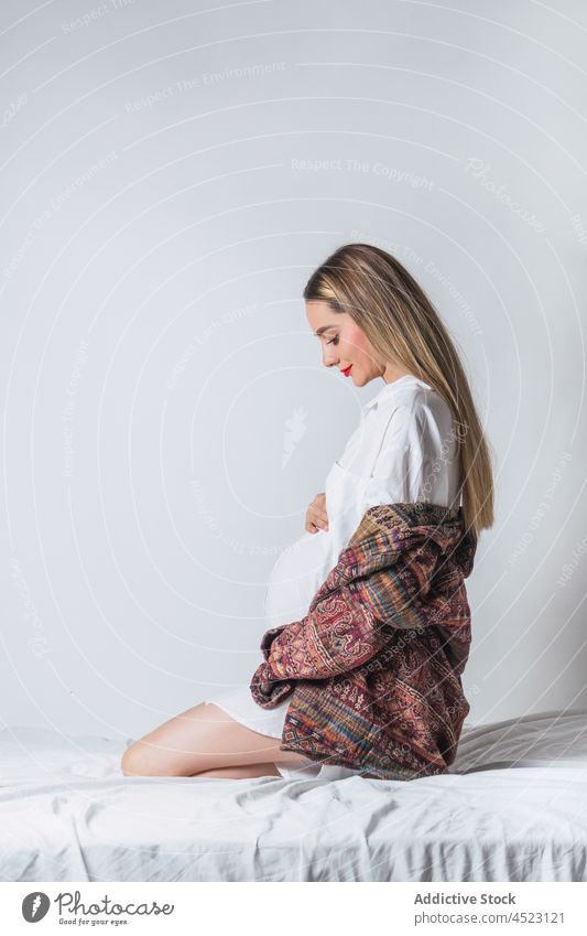 Positive schwangere Frau mit Hand auf dem Bauch auf dem Bett sitzend Angebot sanft mütterlich Harmonie positiv vorwegnehmen erwarten Schwangerschaft sorgenfrei