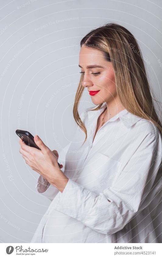 Lächelnde schwangere Frau mit roten Lippen beim Surfen auf dem Smartphone benutzend erwarten positiv Nachricht Internet online vorwegnehmen pränatal
